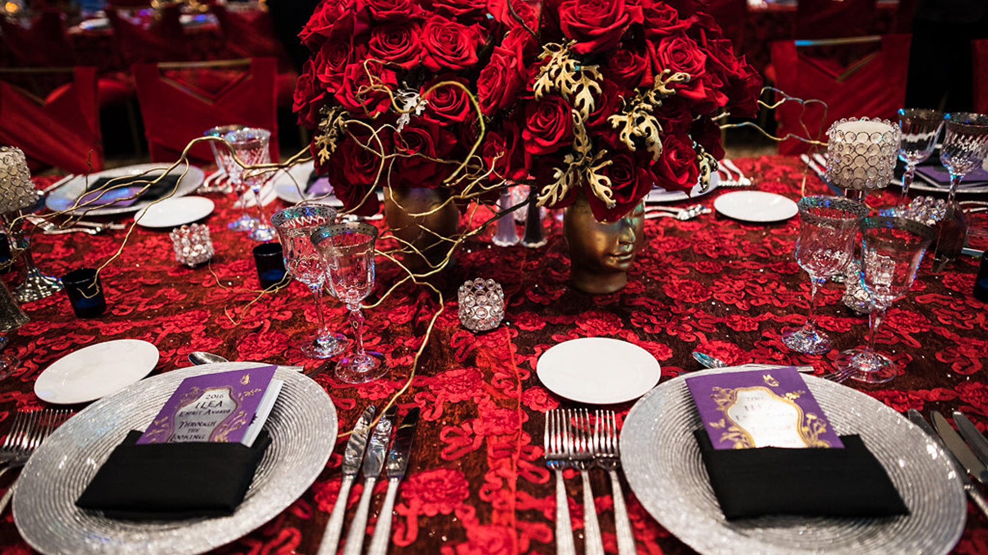 Fancy eating decor atop red rose tablecloth and in front of red roses held in golden-colored vases with the shape of human heads